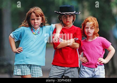 Three children stand side by side outdoors. Two girls with long hair wear casual clothes while a boy in the middle has a serious expression. They show Stock Photo