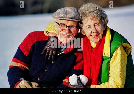 An elderly couple enjoys a fun moment in the snow, smiling and posing for the camera. They hold snowballs while dressed for winter. The sun shines in Stock Photo