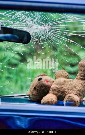 A child's teddy bear lies on the front seat of a blue car. The windshield is cracked and broken after a crash Stock Photo