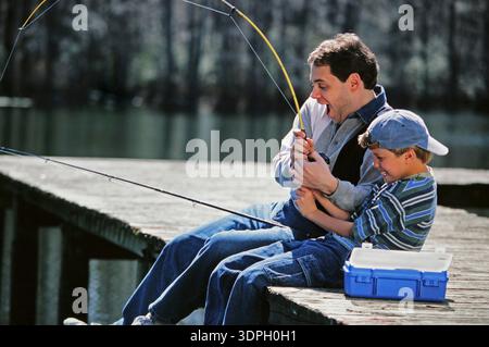 Father and son sit on a wooden dock, working together to reel in a fish. They both show excitement and joy as they enjoy their fishing trip by the lak Stock Photo
