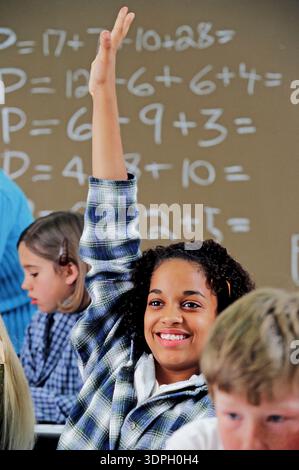Student raises her hand in classroom to answer math question during a lesson on basic arithmetic and problem solving in school Stock Photo