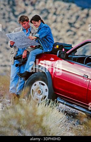 Two people look at a map while sitting on a red car in a desert area during daytime Stock Photo
