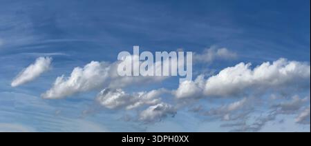 Clouds float in a blue sky on a sunny afternoon. They appear soft and fluffy, moving gently above the landscape. The scene shows a feeling of openness Stock Photo