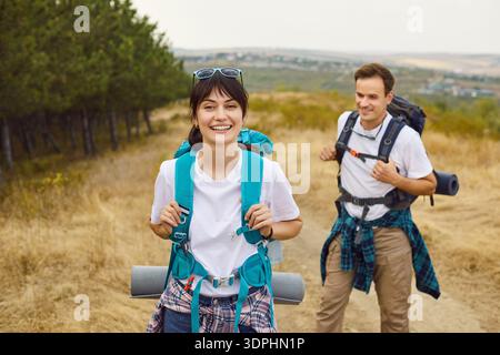 Couple of happy young people hiking on trail by summer forest with backpacks Stock Photo