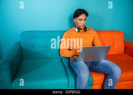 Man sitting on two-tone sofa wearing orange sweatshirt with headphones using laptop, copy space Stock Photo