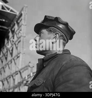 A dramatic archival profile portrait from the 1960s showing a Soviet welder or steelworker at the construction site of the Slavyanskaya Thermal Power Plant (GRES) in Nikolaevka, Donetsk region. The man wears a cap with protective goggles resting on the brim and a sturdy work jacket. He looks resolutely into the distance against a backdrop of a massive steel building skeleton. This authentic close-up captures the strength of the working class and the atmosphere of a peaceful Donbas during the industrial boom. Stock Photo