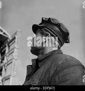 A dramatic archival profile portrait from the 1960s showing a Soviet welder or steelworker at the construction site of the Slavyanskaya Thermal Power Plant (GRES) in Nikolaevka, Donetsk region. The man wears a cap with protective goggles resting on the brim and a sturdy work jacket. He looks resolutely into the distance against a backdrop of a massive steel building skeleton. This authentic close-up captures the strength of the working class and the atmosphere of a peaceful Donbas during the industrial boom. Stock Photo