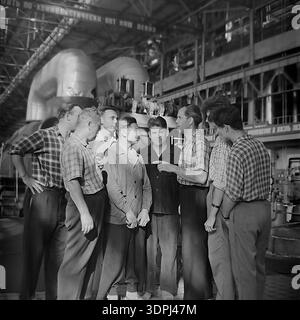 An atmospheric archival photograph from the 1960s capturing a group of Soviet engineers and workers having a lively discussion inside the turbine hall of the Slavyanskaya Thermal Power Plant (GRES) in Nikolaevka, Donetsk region. The men, dressed in period-typical plaid shirts and workwear, stand before massive energy generating equipment. A banner with the slogan "Live and work in a communist way" hangs above. This authentic image illustrates the collective spirit, industrial scale, and the daily life of the peaceful Donbas energy sector. Stock Photo