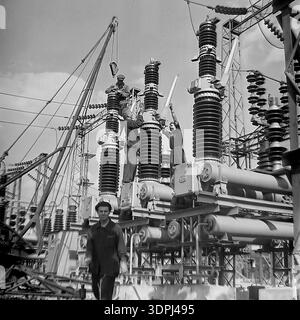 A dramatic vintage archival photograph from the 1960s showing a team of Soviet electricians installing high-voltage circuit breakers at the outdoor substation of the Slavyanskaya Thermal Power Plant (GRES) in Nikolaevka, Donetsk region. Three workers are perched on top of massive porcelain insulators under a crane, while a colleague smiles in the foreground. The image vividly captures the brave work of linemen and the scale of electrical infrastructure in the peaceful Donbas during the Soviet industrial boom. Stock Photo