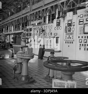 A detailed vintage archival photo from the 1960s showing the interior of the Slavyanskaya Thermal Power Plant (GRES) in Nikolaevka, Donetsk region. In the foreground, large motorized valves with handwheels stand on a tiled floor. In the background, two male operators work at a white control panel: one man in a plaid shirt records data on a clipboard, while his colleague adjusts a switch. This authentic image captures the daily technical routine, teamwork, and industrial scale of the peaceful Donbas energy sector Stock Photo