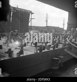 An impressive archival photograph from the 1960s depicting Soviet welders at work on a massive steel grid structure during the construction of the Slavyanskaya Thermal Power Plant (GRES) in Nikolaevka, Donetsk region. The scene captures the intense manual labor and technical skill required to assemble the industrial foundation of the energy giant. Sparks fly as workers in protective masks meticulously weld components together against a backdrop of rising infrastructure. This authentic black and white image represents the creative spirit and the immense engineering scale of the peaceful Donbas Stock Photo
