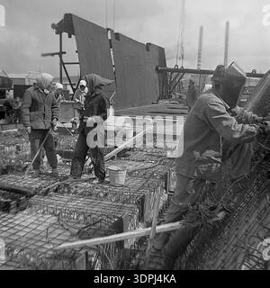 An exceptional archival photograph from the 1960s depicting Soviet construction workers and a welder on a massive rebar grid during the early stages of building the Slavyanskaya Thermal Power Plant in Nikolaevka, Donetsk region. The scene features women in traditional headscarves working alongside technicians amidst complex steel reinforcements, with two tall industrial smokestacks rising in the hazy background. This authentic black and white image perfectly captures the spirit of collective labor, industrial growth, and the peaceful, determined atmosphere of the historic Donbas region. Stock Photo