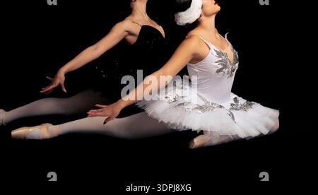 Two ballet dancers perform a synchronized split leap in a studio against a black background. One dancer wears a white classical tutu, the other a blac Stock Photo