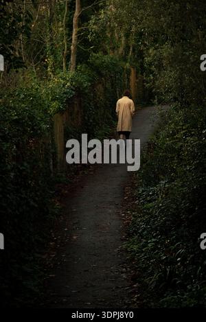 Woman in trench coat walking away on a secluded path in a dense forest. Lonely woman walk in the park Stock Photo