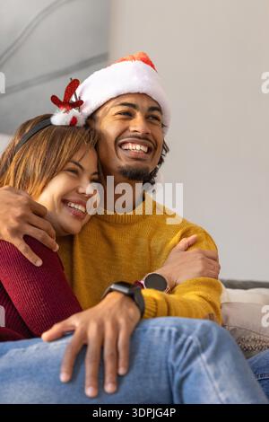 Couple sitting on sofa at home wearing Santa hat and reindeer headband, man wearing smartwatch Stock Photo