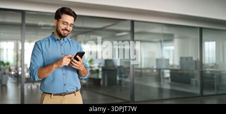 Happy smiling young latin business man holding smartphone standing in office. Stock Photo