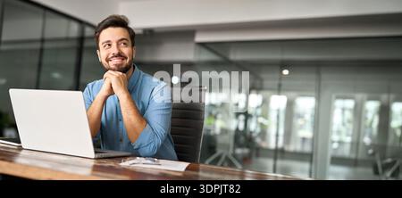 Happy young business man working on laptop looking away at office desk. Stock Photo