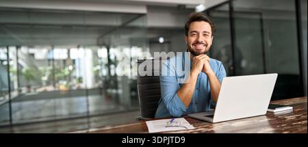 Happy young business man working on laptop looking at camera at office desk. Stock Photo