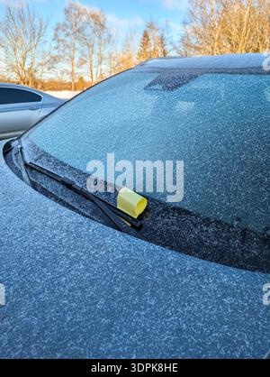 Yellow parking ticket for illegal parking violation placed under the wiper of a frozen car covered in frost during winter. Stock Photo