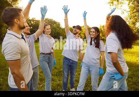 Young happy volunteers standing in a circle in the park holding hands up and smiling Stock Photo