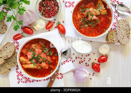Two bowls of Ukrainian borscht on embroidered napkins, bread, sour cream, spices, onions, garlic, tomatoes and parsley on a white wooden background. Horizontal. Top view Stock Photo