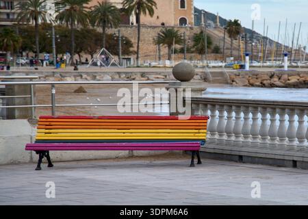 Rainbow painted bench overlooking the seaside promenade and church in Sitges, symbol of diversity, travel destination and Mediterranean lifestyle in S Stock Photo
