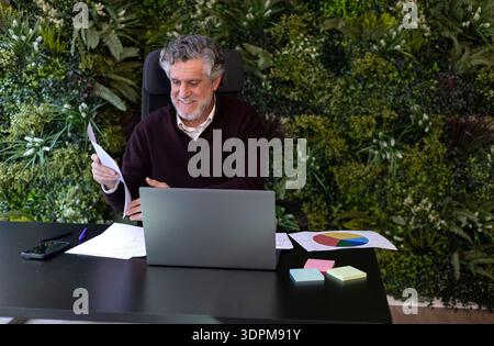 Happy man reading paper while sitting at desk with laptop in home office Stock Photo