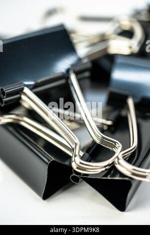 Several black binder clips are stacked and arranged on a flat white surface. They are shiny and metallic, showing their clips blending in with each ot Stock Photo