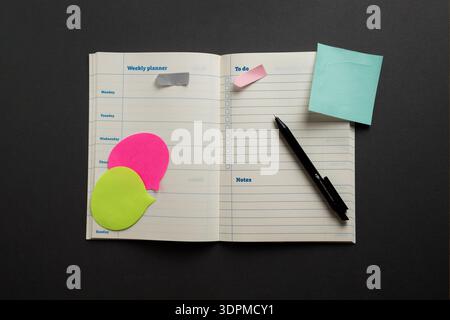 Open weekly planner with colorful sticky notes and pen arranged on dark background, minimalist planning and organization workspace. Stock Photo