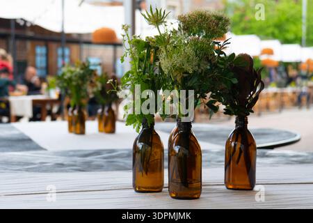 Small delicate bouquets of wild flowers and greenery in amber glass apothecary bottles on a white table in an outdoor summer cafe Stock Photo