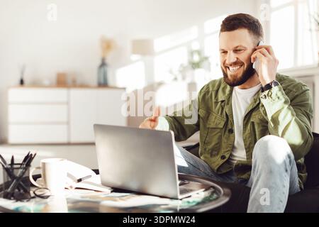 Joyful Freelancer Man Talking On Phone Using Laptop At Home Stock Photo