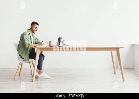 Young Man Studying at a Wooden Desk With Laptop and Headphones in a Bright Room Stock Photo