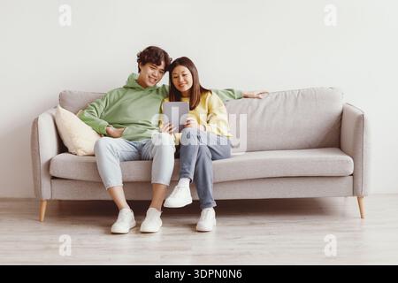 Happy Asian couple sitting on couch with tablet, looking at touch pad screen, using newest application at home Stock Photo