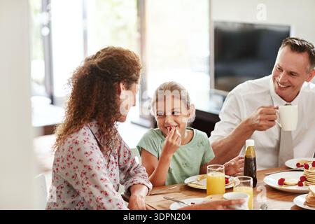 Happy, parents and kid with breakfast in home for morning meal, start day or bonding with nutrition. Family, eating and talking to child at dining Stock Photo