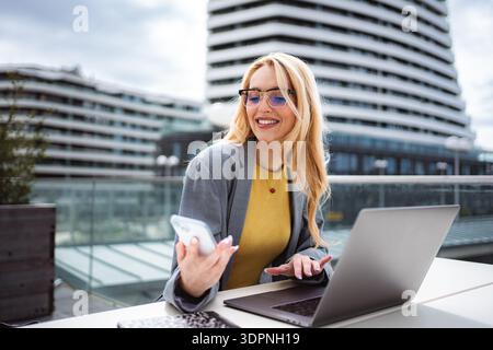 Woman smiling while using phone and laptop on a city patio Stock Photo