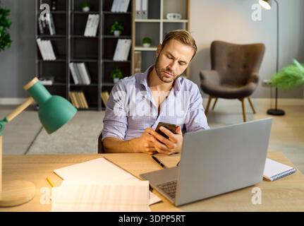 Busy sad man using phone and laptop, suffering from overtime work at office Stock Photo