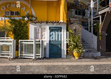 Parga, Greece - October 26, 2020: A traditional blue wooden door and white-framed window are set in a yellow building facade, with a potted flowering plant, stone steps leading upward, and a small stone shrine visible beside the entrance. Stock Photo
