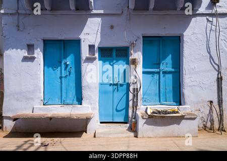 Udaipur, India - March 23, 2022: Three closed blue wooden doors are set in a whitewashed wall with two concrete benches and an electrical meter with exposed wiring visible on a sunny street. Stock Photo
