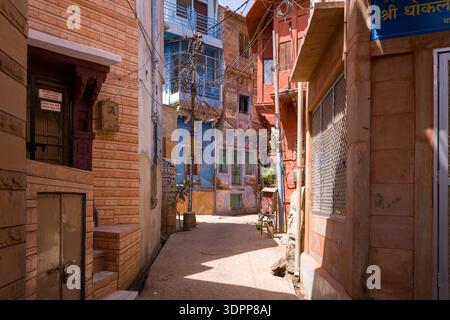 Jodhpur, India - March 26, 2022: A narrow alleyway is lined with blue, red, and tan residential buildings featuring barred windows, signage in Hindi, and numerous overhead electrical wires under bright daylight. Stock Photo