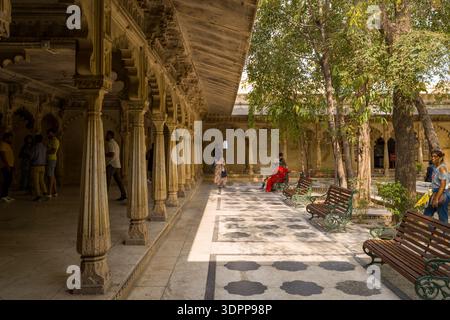 Udaipur, India - March 24, 2022: Several people are present in the tree-lined inner courtyard of City Palace, with stone benches, patterned paving, and arched corridors visible under a covered walkway. Stock Photo