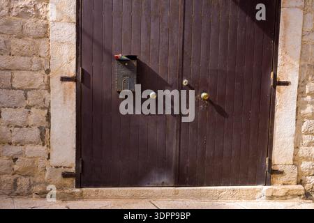 Trani, Italy - October 16, 2020: A metal mailbox labeled 'Posta' with a rolled newspaper sticking out is mounted on a brown wooden door with two round handles, next to a stone wall, seen in sunlight. Stock Photo