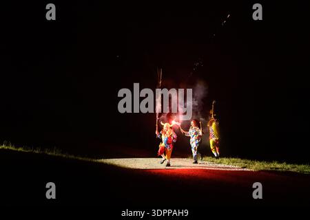 Unknown location - July 17, 2021: Three people dressed in colorful costumes are holding fireworks that emit sparks and smoke into the night sky during an outdoor performance on a grassy field. Stock Photo