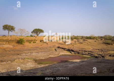Chittorgarh, India - March 21, 2022: A dry reservoir with exposed rocky ground is seen in front of a stone fort wall topped with trees and a small domed pavilion at Maharani Padmini Reservoir, with sparse vegetation and clear sky in the background. Stock Photo