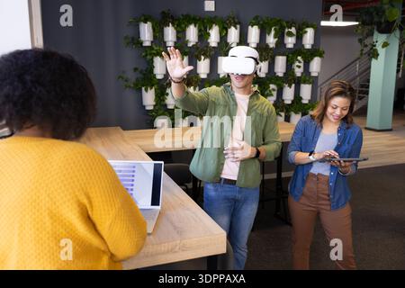 Diverse coworkers in office using VR headset, working on tablet and laptop by vertical garden wall Stock Photo