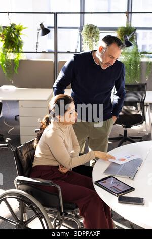 Diverse coworkers analyzing printed data report using wheelchair and laptop at modern office Stock Photo