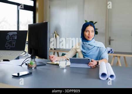 Indian Asian woman sitting at desk in workspace reviewing blueprints on dual monitors with laptop Stock Photo