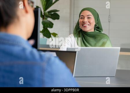 Diverse female coworkers smiling and collaborating on laptops at office desk with plant, copy space Stock Photo