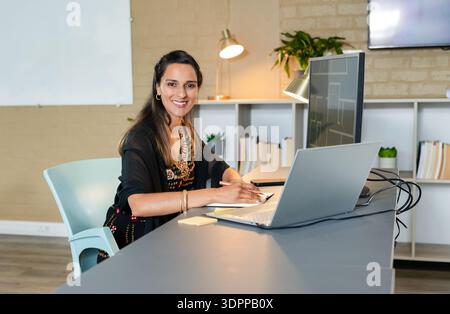 Indian adult woman smiling and writing notes on notepad at desk with laptop floor plan monitor Stock Photo