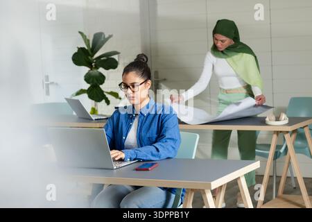 Diverse female colleagues collaborating in bright office using laptop and studying blueprints Stock Photo