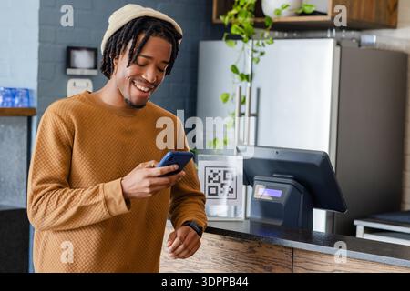 African American man scanning acrylic QR code using smartphone at cafe counter with POS screen Stock Photo
